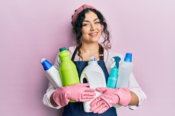 Young brunette woman with curly hair wearing cleaner apron holding cleaning products smiling with a...