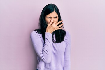 Young hispanic woman wearing casual clothes smelling something stinky and disgusting, intolerable smell, holding breath with fingers on nose. bad smell