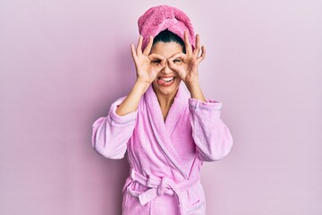 Young hispanic woman wearing shower towel cap and bathrobe doing ok gesture like binoculars sticking tongue out, eyes looking through fingers. crazy expression.