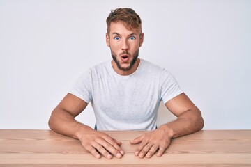 Young caucasian man wearing casual clothes sitting on the table scared and amazed with open mouth for surprise, disbelief face