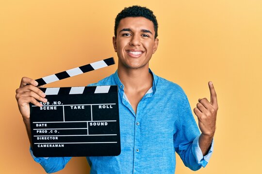 Young arab man holding video film clapboard smiling with an idea or question pointing finger with happy face, number one