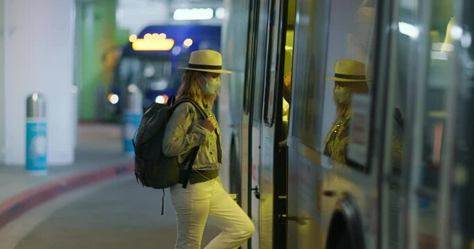 Woman in protective face mask boarding the bus shuttle at the arrival or departure area of the international airport terminal. Traveler with backpack getting on the bus. COVID-19 pandemic, 4K USA