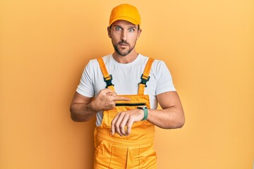 Young handsome man wearing handyman uniform over yellow background in hurry pointing to watch time, impatience, upset and angry for deadline delay