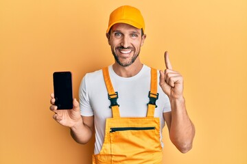 Young handsome man wearing handyman uniform showing smartphone screen smiling with an idea or...