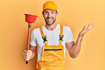 Young handsome man wearing plumber uniform holding toilet plunger celebrating achievement with...