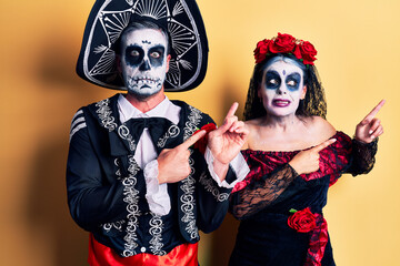 Young couple wearing mexican day of the dead costume over yellow pointing aside worried and nervous with both hands, concerned and surprised expression
