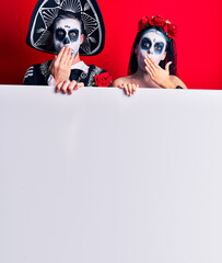 Young couple wearing mexican day of the dead costume holding blank empty banner covering mouth with hand, shocked and afraid for mistake. surprised expression