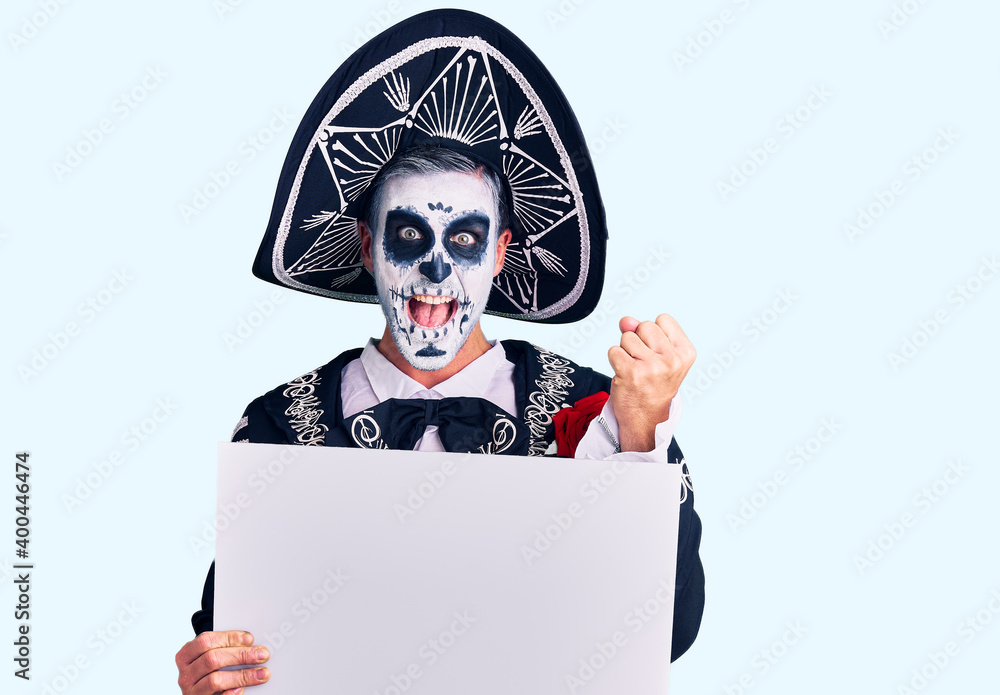 Wall mural Young man wearing day of the dead costume holding blank empty banner screaming proud, celebrating victory and success very excited with raised arms