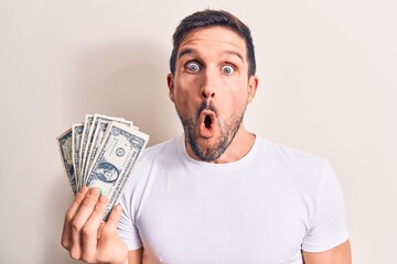 Young handsome man holding bunch of dollars banknotes over isolated white background scared and amazed with open mouth for surprise, disbelief face