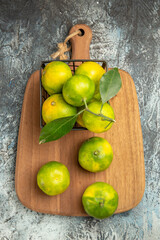 Overhead view of green mandarins with leaves inside and outside of a basket on wooden cutting board on gray table