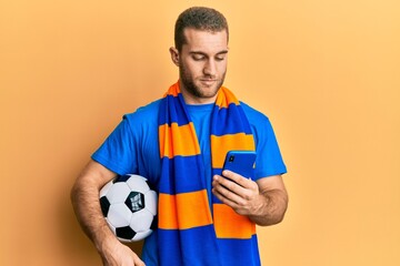 Young caucasian man holding football ball looking at smartphone relaxed with serious expression on face. simple and natural looking at the camera.