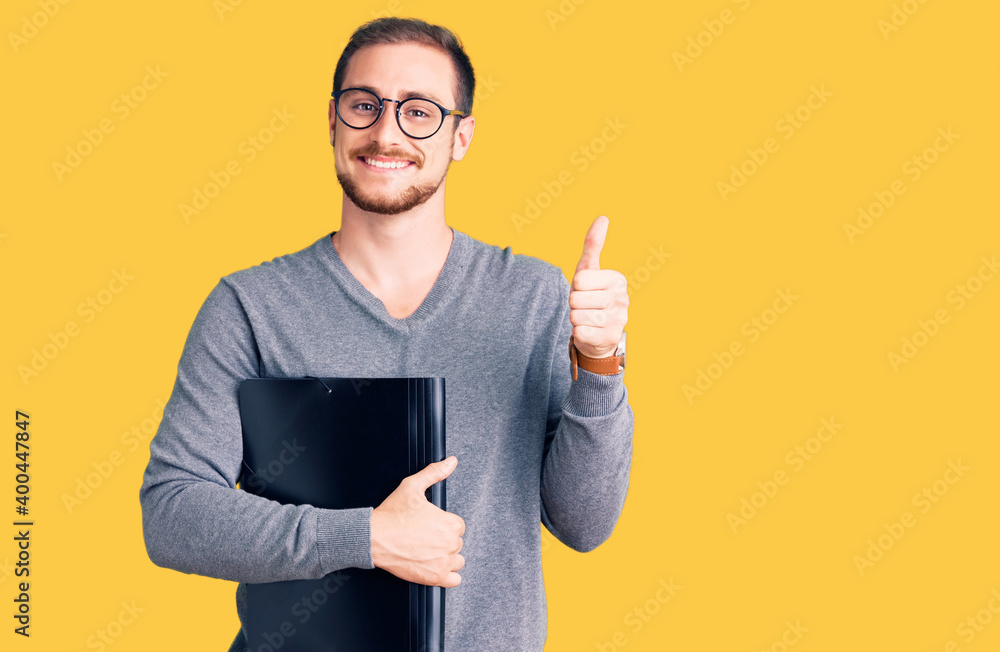 Wall mural young handsome caucasian man holding business folder smiling happy and positive, thumb up doing exce