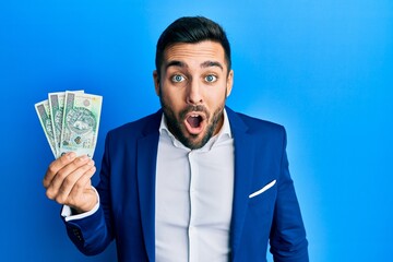 Young hispanic businessman wearing business suit holding polish zloty banknotes scared and amazed with open mouth for surprise, disbelief face