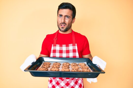 Young hispanic man wearing baker uniform holding homemade cookies clueless and confused expression. doubt concept.