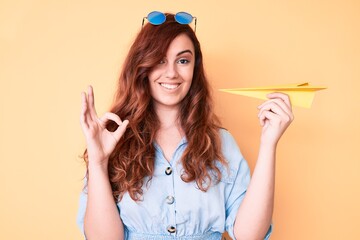 Young beautiful woman holding paper airplane doing ok sign with fingers, smiling friendly gesturing excellent symbol