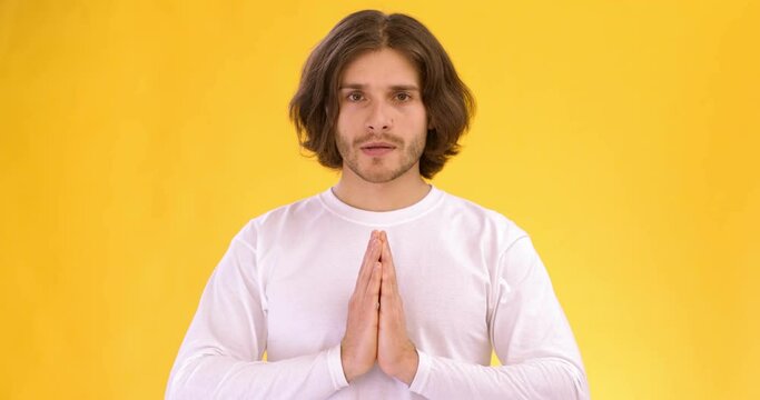 Studio portrait of peaceful calm inspired man ib white shirt holding hands in prayer gesture, orange background