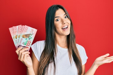 Young brunette woman holding 100 new zealand dollars banknotes celebrating achievement with happy smile and winner expression with raised hand