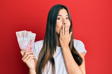 Young brunette woman holding 500 swedish krona banknotes covering mouth with hand, shocked and afraid for mistake. surprised expression