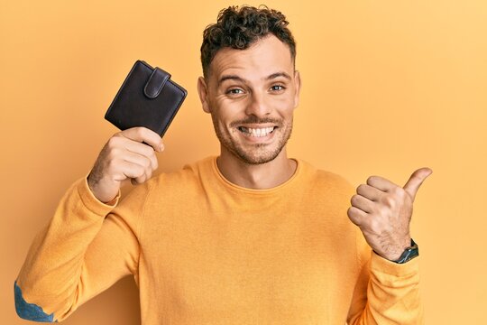 Young hispanic man holding leather wallet pointing thumb up to the side smiling happy with open mouth