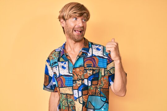 Young blond man wearing summer shirt smiling with happy face looking and pointing to the side with thumb up.