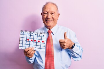 Senior handsome grey-haired businessman wearing tie holding cardio heart calendar smiling happy and positive, thumb up doing excellent and approval sign