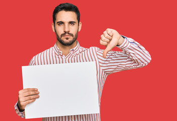 Young hispanic man holding blank empty banner with angry face, negative sign showing dislike with thumbs down, rejection concept