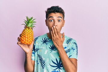 Young handsome african american man holding pineapple covering mouth with hand, shocked and afraid...