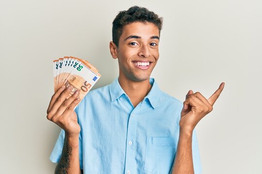 Young handsome african american man holding bunch of 50 euro banknotes smiling happy pointing with hand and finger to the side