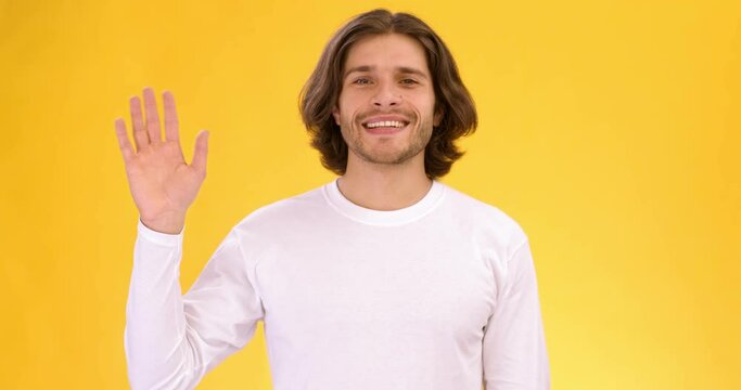 Studio shot of young friendly talkative guy waving hand and saying Hello, slow motion, orange background