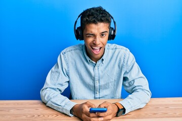 Young handsome african american man using smartphone wearing headphones winking looking at the...