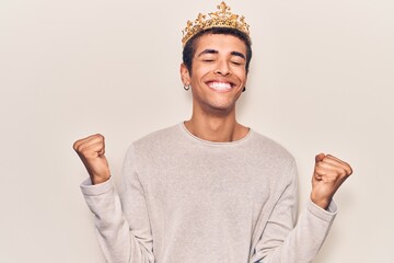 Young african amercian man wearing prince crown very happy and excited doing winner gesture with...