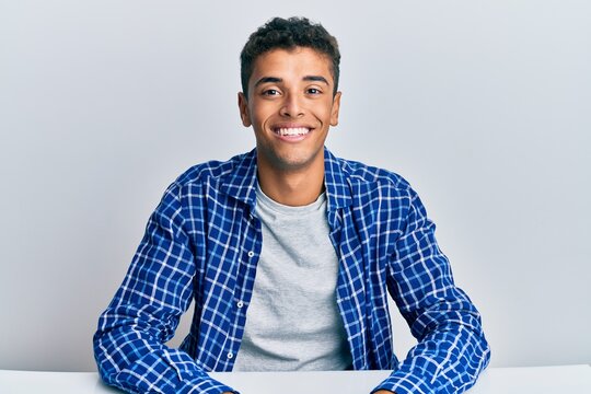 Young handsome african american man wearing casual clothes sitting on the table looking positive and happy standing and smiling with a confident smile showing teeth