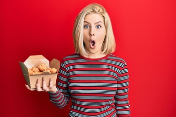 Young blonde woman eating fried chicken scared and amazed with open mouth for surprise, disbelief...