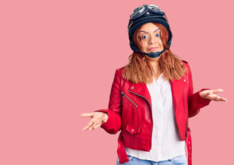 Young latin woman wearing leather jacket holding motorcycle helmet clueless and confused expression with arms and hands raised. doubt concept.