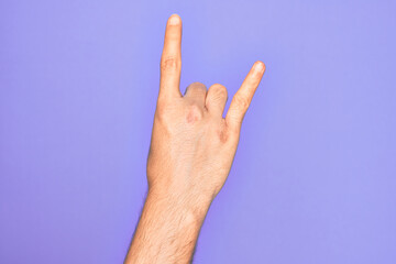 Hand of caucasian young man showing fingers over isolated purple background gesturing rock and roll symbol, showing obscene horns gesture