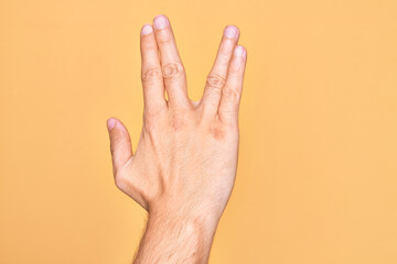 Hand of caucasian young man showing fingers over isolated yellow background greeting doing Vulcan salute, showing back of the hand and fingers, freak culture