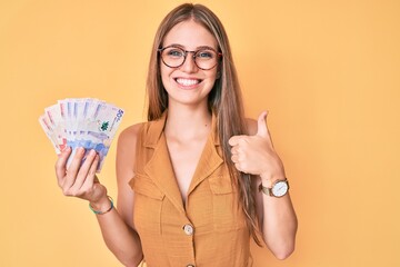 Young blonde girl holding colombian pesos smiling happy and positive, thumb up doing excellent and approval sign