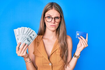 Young blonde girl holding credit car and dollars skeptic and nervous, frowning upset because of...