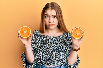 Young beautiful redhead woman holding fresh orange slices skeptic and nervous, frowning upset...