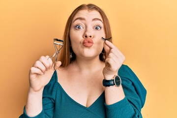 Young beautiful redhead woman holding eyelash curler and fake lashes looking at the camera blowing a kiss being lovely and sexy. love expression.