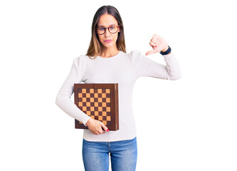 Beautiful brunette young woman holding chess with angry face, negative sign showing dislike with...