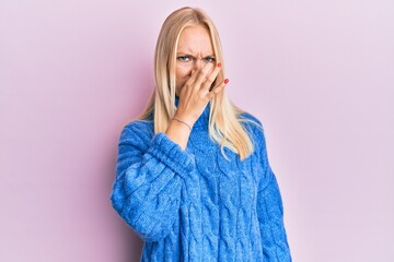 Young blonde girl wearing wool winter sweater smelling something stinky and disgusting, intolerable smell, holding breath with fingers on nose. bad smell