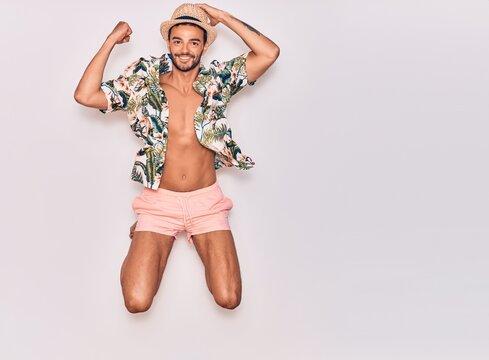 Young handsome hispanic man on vacation wearing swimwear, floral shirt and hat smiling happy. Jumping with smile on face celebrating with fist up over isolated white background