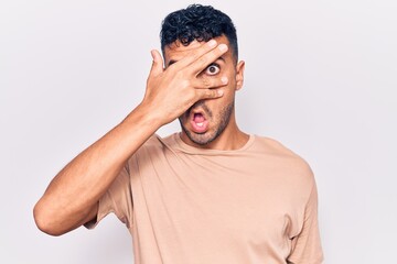 Young hispanic man wearing casual clothes peeking in shock covering face and eyes with hand,...