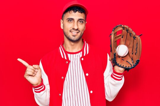 Young hispanic man wearing baseball uniform holding golve and ball smiling happy pointing with hand and finger to the side