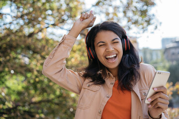 Young beautiful African American woman listening to music on headphone, dancing, singing songs. Emotional female holding mobile phone, celebration success 