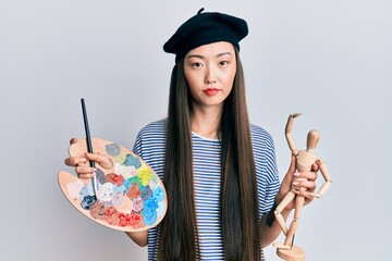 Young chinese woman wearing artist look with beret holding manikin relaxed with serious expression on face. simple and natural looking at the camera.