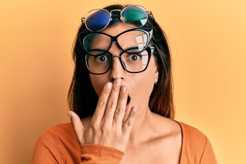 Beautiful hispanic woman wearing many glass over face covering mouth with hand, shocked and afraid...