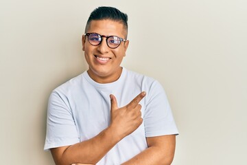 Young latin man wearing casual clothes and glasses smiling cheerful pointing with hand and finger up to the side