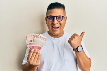 Young latin man holding 10 colombian pesos banknotes pointing thumb up to the side smiling happy with open mouth